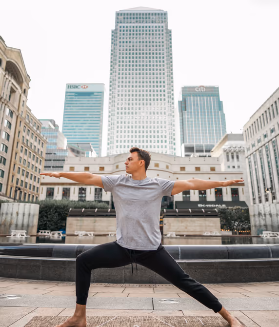 A teacher in Warrior II pose on a yoga mat outdoors, with the Canary Wharf skyline behind.